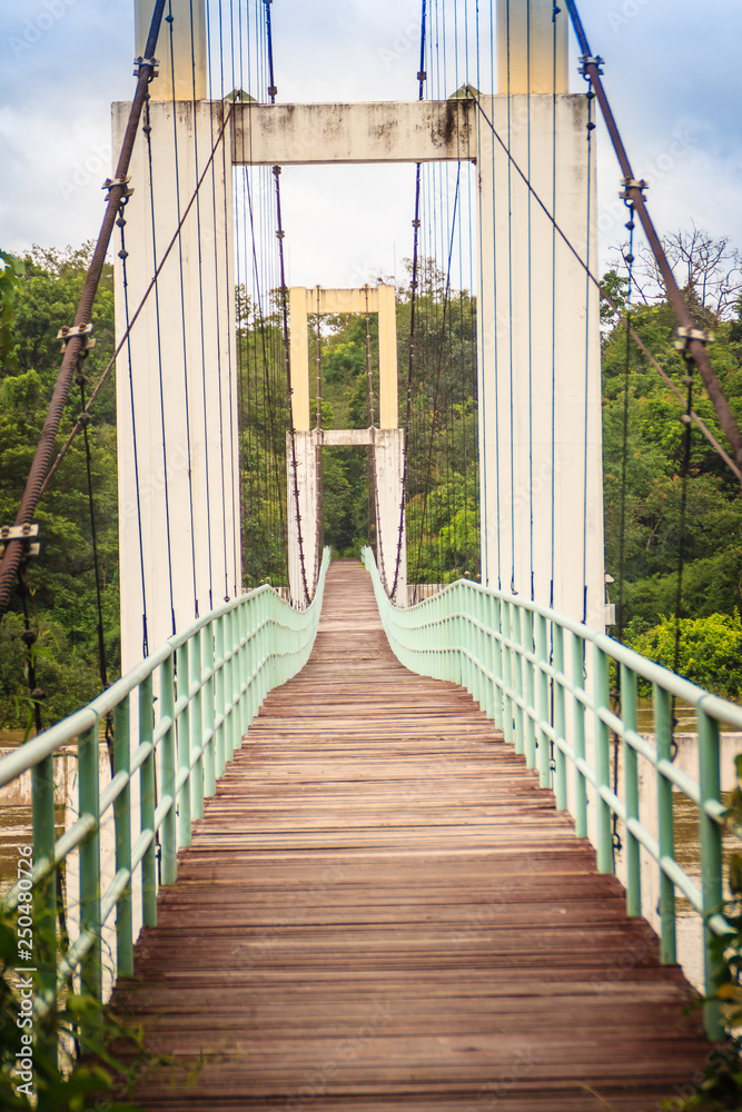 Obraz premium Vintage suspension bridge hanging across the river. Old lengthy hanging wooden footbridge with steel rails over river against cloudy sky background. Hanging wooden bridge at Kaengtana national park.