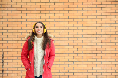 Beautiful young woman listening to music with headphones against brick wall. Space for text