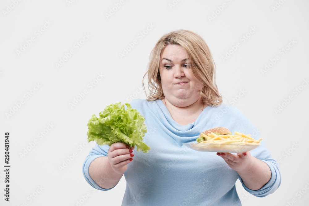 Young fluffy fat woman in casual blue clothes on a white background holding green salad leaves and a plate of fast food, hamburger and fries. Diet and proper nutrition. Young fluffy fat woman in casual blue clothes on a white background holding green salad leaves and a plate of fast food, hamburger and fries. Diet and proper nutrition.