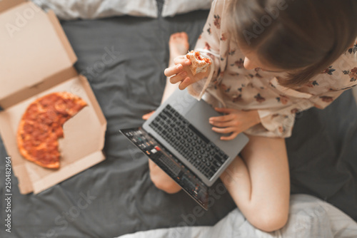 Portrait of a girl sitting in a bed with a box of pizza and using the Internet on a laptop, holds a piece of pizza in her hand. Work and breakfast in bed. Fast Food Concept. Pizza delivery.