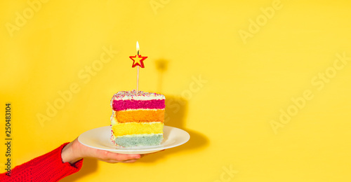 Female hand holding white plate with slice of Rainbow cake with birning candle in the shape of star isolated on yellow background Fototapete