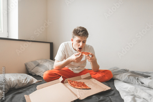 Portrait of a young man sitting at home in bed and eating pizza from a box holding a piece of pizza in his hands.Guy is eating fast food from the delivery.Teen eats a pizza from the box in the bedroom