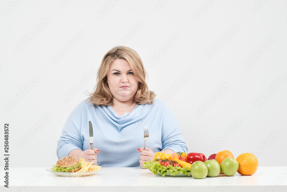 Young curvy fat woman in casual blue clothes on a white background at the table chooses what to eat: fast food or healthy food. Diet and proper nutrition. Young curvy fat woman in casual blue clothes on a white background at the table chooses what to eat: fast food or healthy food. Diet and proper nutrition.