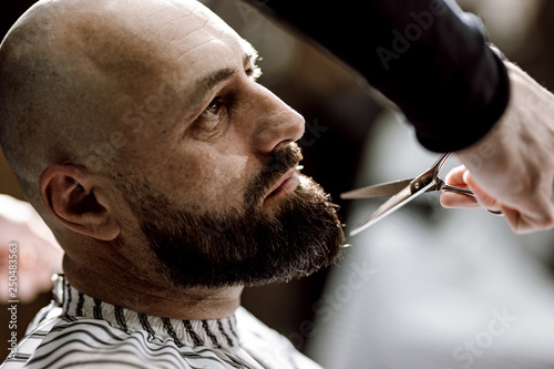 Fashion barber dressed in a black clothes scissors beard of brutal man in the stylish barbershop