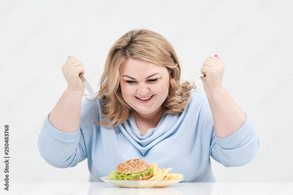 Young curvy fat woman in casual blue clothes on a white background at the table eagerly eating fast food, hamburger and french fries. Diet and proper nutrition. Young curvy fat woman in casual blue clothes on a white background at the table eagerly eating fast food, hamburger and french fries. Diet and proper nutrition.