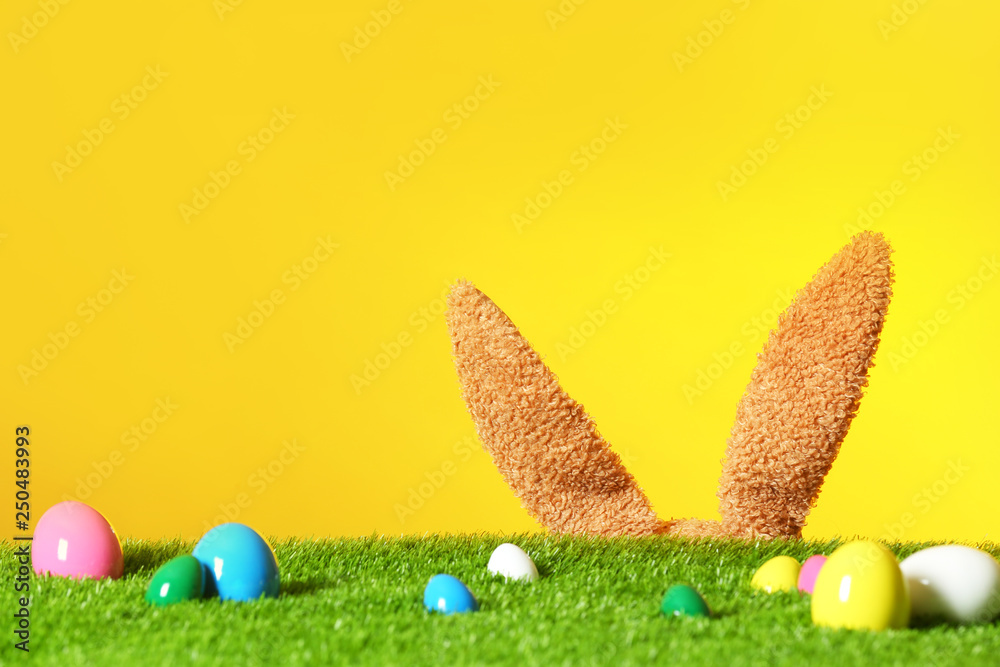 Easter eggs on green lawn and funny bunny ears against color background, space for text