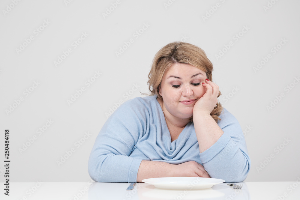 Young curvy fat woman in casual blue clothes on a white background at the table sitting in front of an empty plate and starving. Diet and proper nutrition. Young curvy fat woman in casual blue clothes on a white background at the table sitting in front of an empty plate and starving. Diet and proper nutrition.