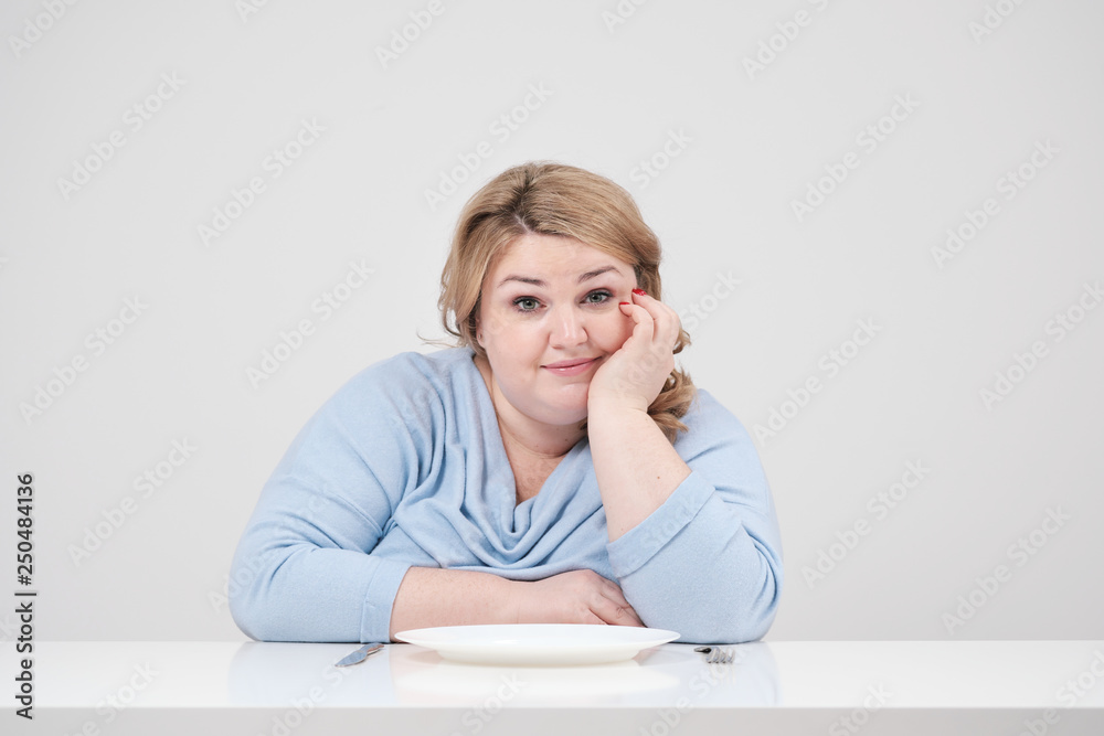 Young curvy fat woman in casual blue clothes on a white background at the table sitting in front of an empty plate and starving. Diet and proper nutrition. Young curvy fat woman in casual blue clothes on a white background at the table sitting in front of an empty plate and starving. Diet and proper nutrition.