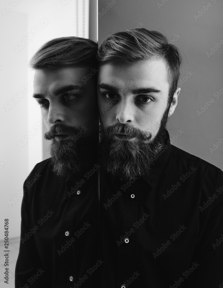 Obraz premium Black and white photo of a man with a beard and stylish hairdo dressed in the black shirt standing next to the mirror with his reflection