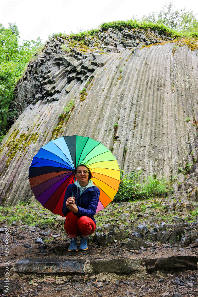 Young woman in glasses sit under the colorful umbrella with famous somoska stone waterfall on the background, Slovakia