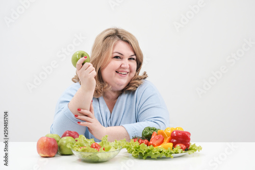 Young curvy fat woman in casual blue clothes on a white background at the table and shows OK, vegetables and fruits are laid out in front of her in a row. Diet and proper nutrition.