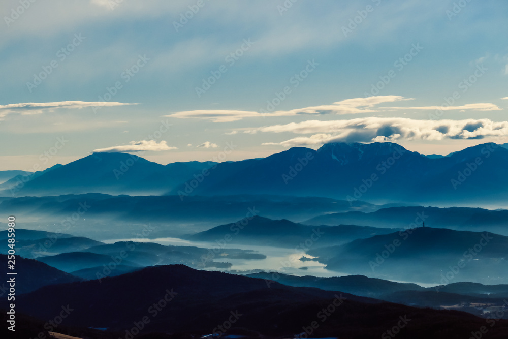 View from Dobratsch on Valley of Roses, Austria. Valley is covered with morning for, created by Wörthersee Lake. Few clouds above the Alps. Serene and tender morning. Soft light. Delicate colors.