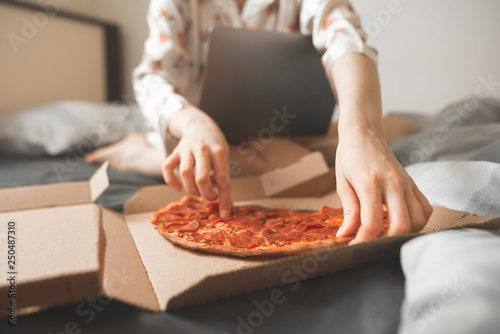 Women's hands take a piece of pizza from a box, sitting on a bed using a laptop. Pizza in bed, fast food. Focus on the pizza. Background. Copyspace © bodnarphoto