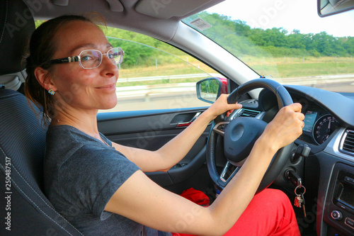 Happy smiling young woman in glasses driving a car with green trees ove rthe window
