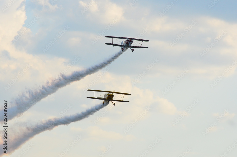 Fototapeta premium Two stunt biplanes flying one above the other on a cloudy blue sky leaving white smoke trail