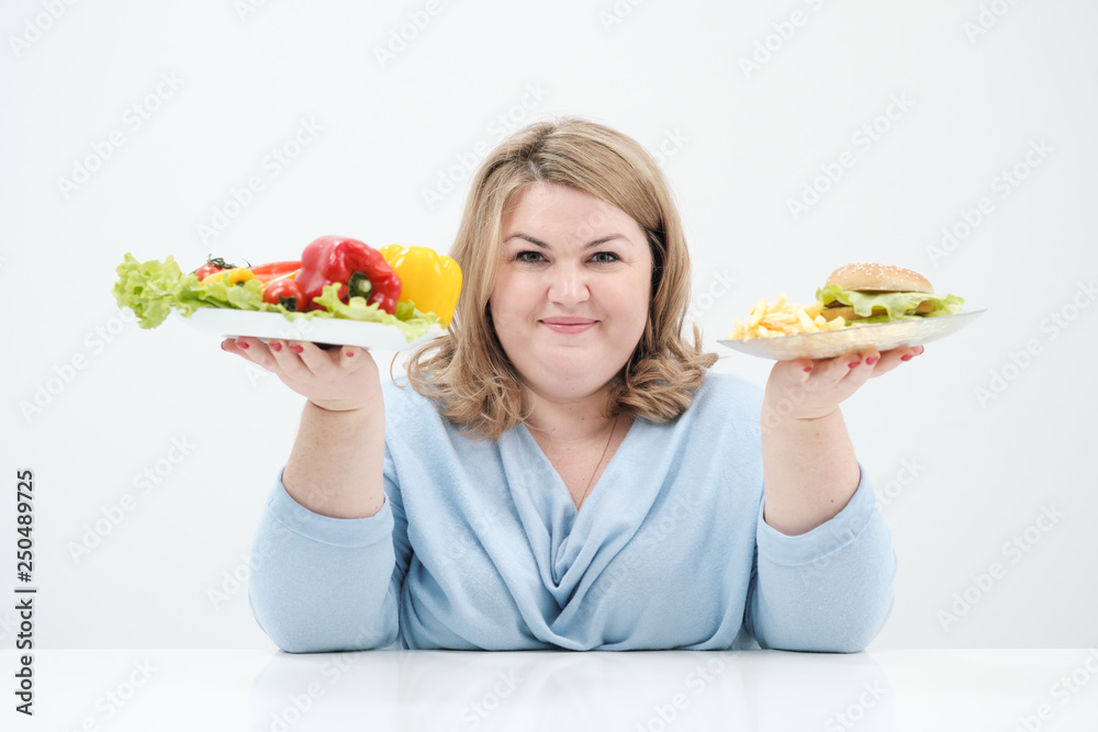 Young lush fat woman in casual blue clothes on a white background holding a vegetable salad and a plate of fast food, hamburger and fries. Diet and proper nutrition. Young lush fat woman in casual blue clothes on a white background holding a vegetable salad and a plate of fast food, hamburger and fries. Diet and proper nutrition.
