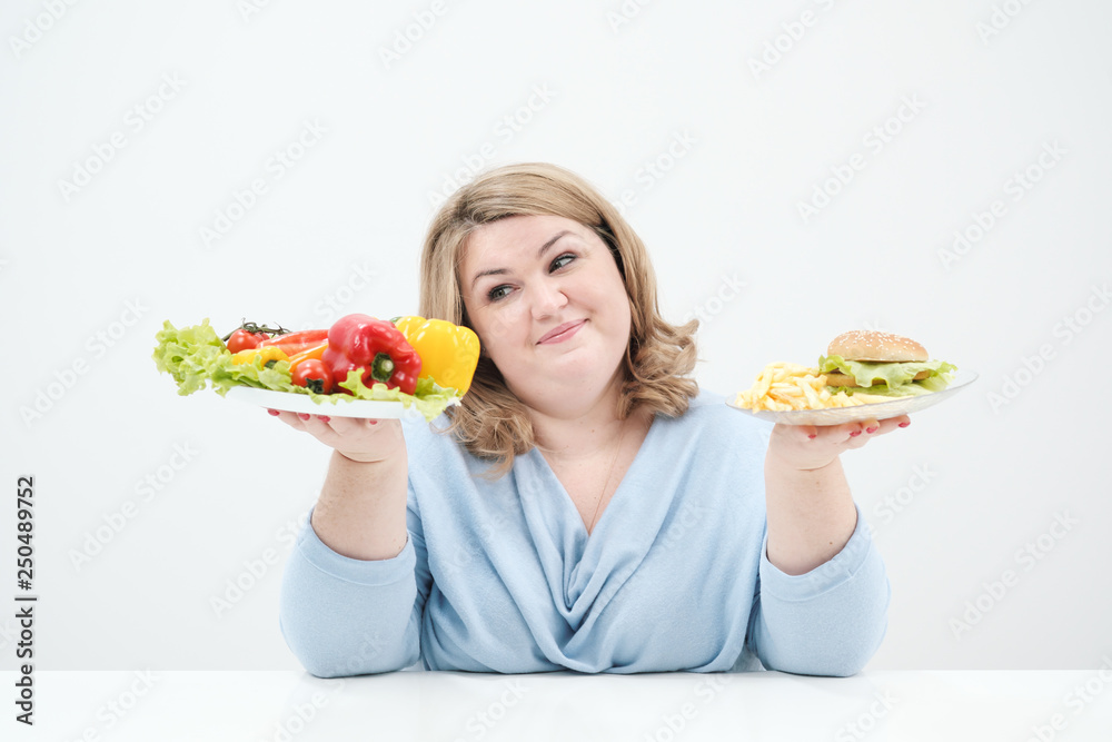 Young lush fat woman in casual blue clothes on a white background holding a vegetable salad and a plate of fast food, hamburger and fries. Diet and proper nutrition. Young lush fat woman in casual blue clothes on a white background holding a vegetable salad and a plate of fast food, hamburger and fries. Diet and proper nutrition.