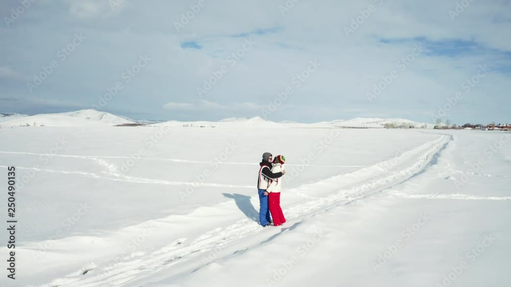 Aerial drone view; camera moves around the young couple hugging on the frozen lake among the pure white snow in bright clothes; enjoying of winter nature with mounts on background; love and romantic Aerial drone view; camera moves around the young couple hugging on the frozen lake among the pure white snow in bright clothes; enjoying of winter nature with mounts on background; love and romantic