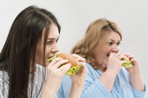 Two girls  slim and fat  blonde and brunette  eat hamburgers. On a white background  the theme of diet and proper nutrition.