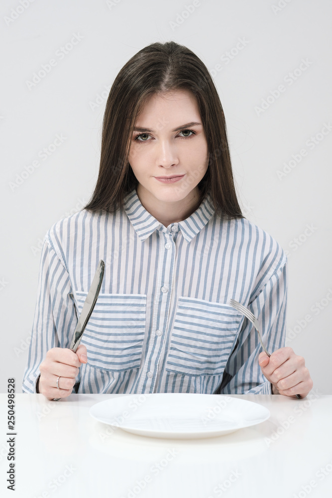 Hungry thin girl sitting at the table in front of an empty plate with a knife and fork. Hungry thin girl sitting at the table in front of an empty plate with a knife and fork.