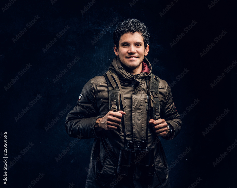 Smiling young traveller in hiking gear with binoculars looking at a camera while standing in the studio against a dark wall Smiling young traveller in hiking gear with binoculars looking at a camera while standing in the studio against a dark wall