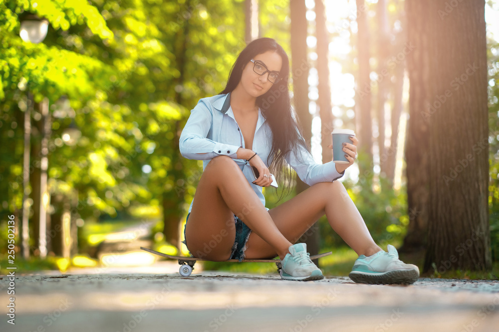 A beautiful girl in a white shirt and shorts holding a smartphone and cup of takeaway coffee while sitting on a skateboard in the park on a summer sunny day.