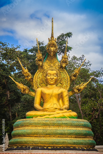 Beautiful golden Buddha statue with seven Phaya Naga heads under white clouds and blue sky background. Outdoor golden seated Buddha image protected by 7 heads Naga spreads cover on top in cloudy days.