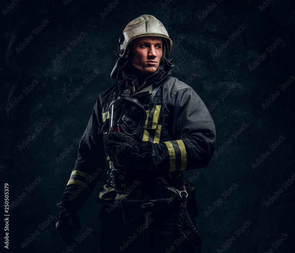 Brutal firefighter in uniform and safety helmet holding an oxygen mask and looking sideways with a confident look. Studio portrait in a dark studio against a textured wall. Brutal firefighter in uniform and safety helmet holding an oxygen mask and looking sideways with a confident look. Studio portrait in a dark studio against a textured wall.