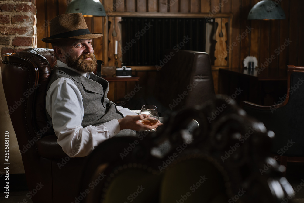 Φωτογραφία A man in hat wearing vintage suit holding pipe and glass of whiskey sitting on a