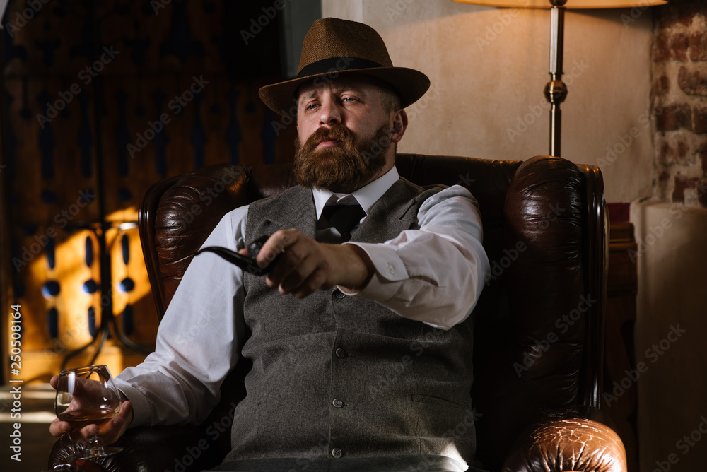 Φωτογραφία A man in hat wearing vintage suit holding pipe and glass of whiskey sitting on a