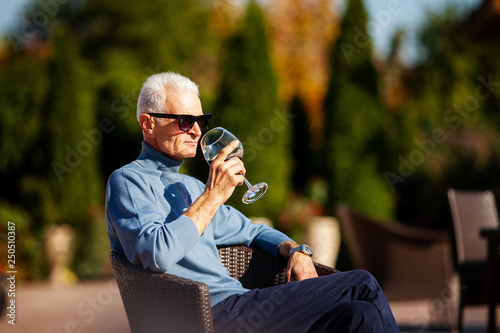 Relaxed old grey-haired man sit at the garden and enjoy glass of wine