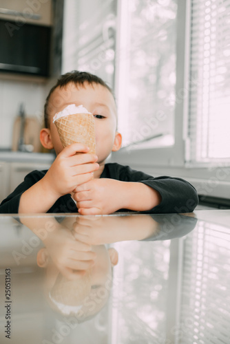 A child in a dark-blue t-shirt in the bright kitchen eating a waffle ice cream cone in the summer house photo