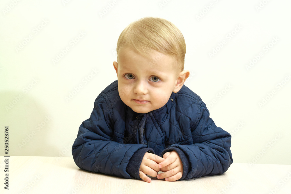 little bright-haired pretty boy sitting behind a light table in a blue jacket hands holding together little bright-haired pretty boy sitting behind a light table in a blue jacket hands holding together