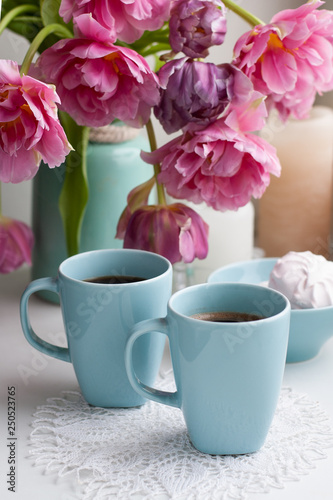 Two cups of black coffee and a delicate marshmallow for breakfast are next to a beautiful bouquet of pink tulips in a blue vase.