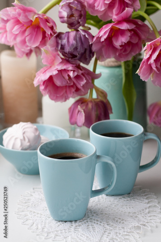 Two cups of black coffee and a delicate marshmallow for breakfast are next to a beautiful bouquet of pink tulips in a blue vase.