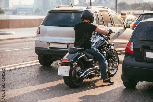 rear view of a handsome biker dressed in a black T-shirt and black helmet jeans and sneakers. He is riding on cruiser motorcycle on background of 2 cars. Traffic stop