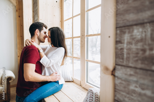 Beautiful guy and girl in stylish clothes hug each other. The girl sits on the windowsill, the guy stands next to her © Kateryna