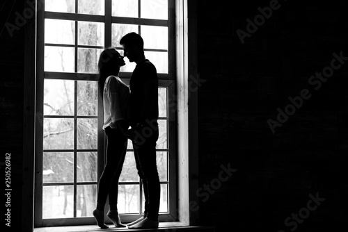 Silhouette of a couple that stands near a large window, looks at each other and holds hands. Black and white photo