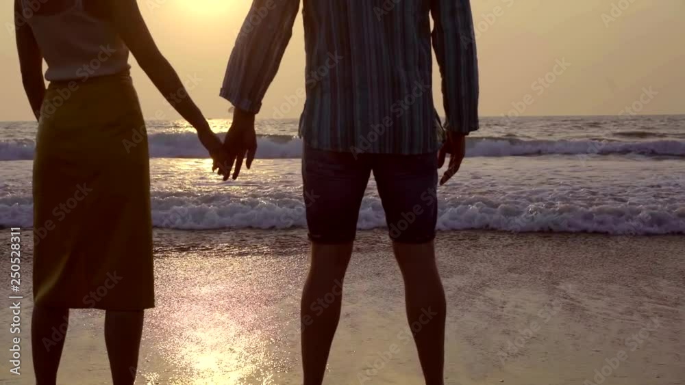 Shot from behind of a couple in love standing on the beach and looking at sun going down.