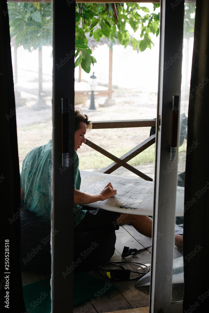 Young man rubbing wax on surfboard while sitting at porch seen through doorway Young man rubbing wax on surfboard while sitting at porch seen through doorway