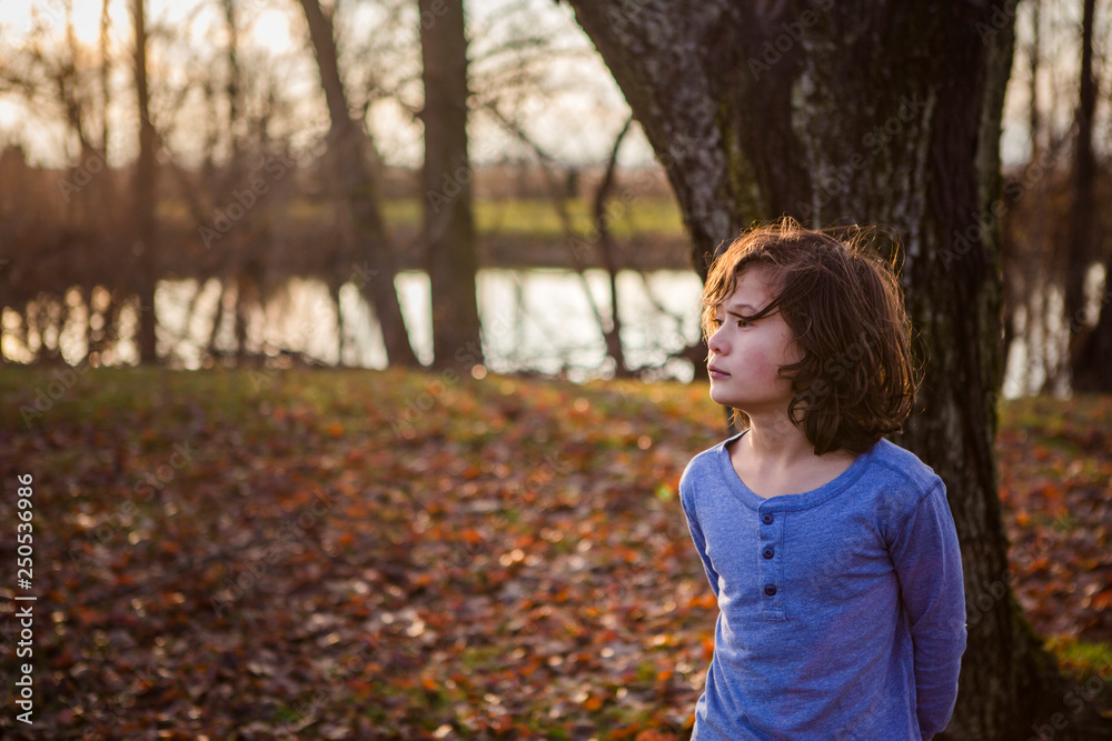 Thoughtful boy standing near tree trunk in park during sunset