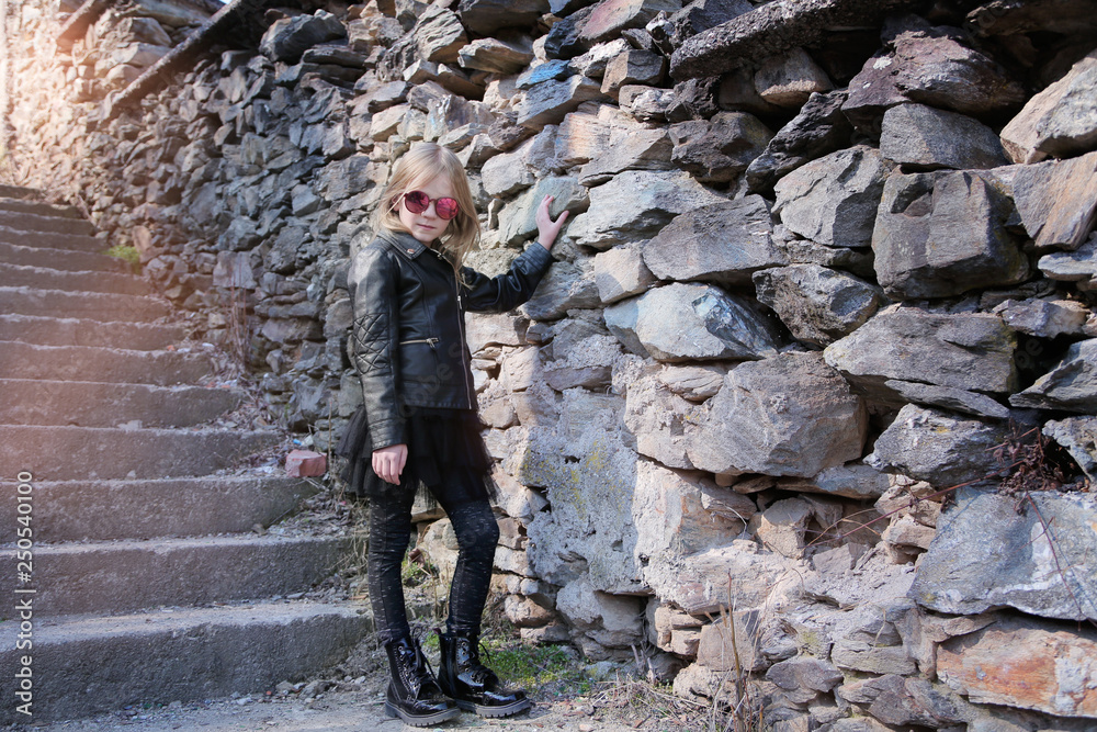 Beautiful blond little girl, dressed in rock and roll style: black leather jacket, skirt and boots sunglasses. The girl stay on a ladder near a stone wall.