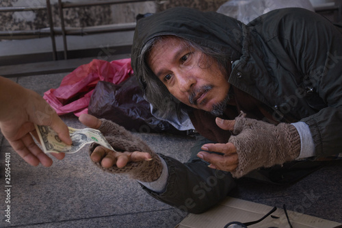 Homeless man is sitting down on walkway in town. He is receive dollar.He is hoping to have the better life. poverty, despair, Photo Sympathetic and hope concept.