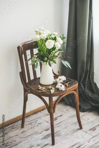 Spring white buttercup flowers in enamel jug on wooden vintage chair on floor with white wall at background. Wedding bouquet, flower shop, Women's Day holiday or spring mood concept photo