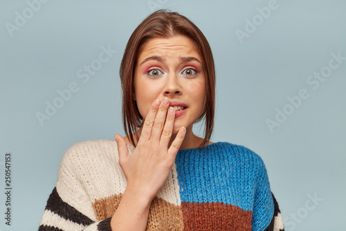 Confused young girl shrugging shoulders, having guilty look, feeling sorry for doing something wrong and making terrible mistake, keeps hand on lips, over blue background photo