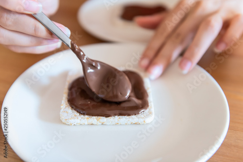 Closeup of rice cake with woman hand spreading chocolate hazelnut spread with spoon sauce or syrup brown vegan vegetarian snack dessert one single piece