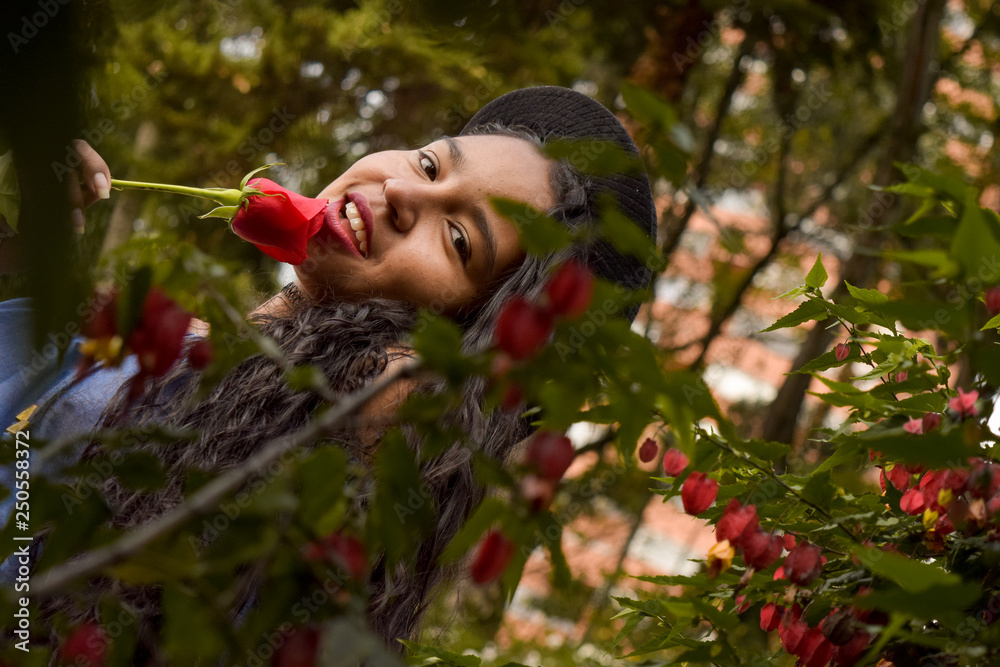 Fototapeta premium Beautiful and seductive young woman with hat, passionately holds a rose in her mouth. She looks at us with madness through a foliage of leaves