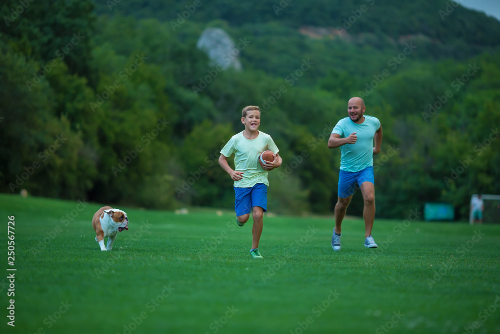 Father and son are running in park with dog english bulldog. Father's Day. Father with son and dog on a walk in the city park on sunny summer day. Family walking with dog in park Father and son are running in park with dog english bulldog. Father's Day. Father with son and dog on a walk in the city park on sunny summer day. Family walking with dog in park