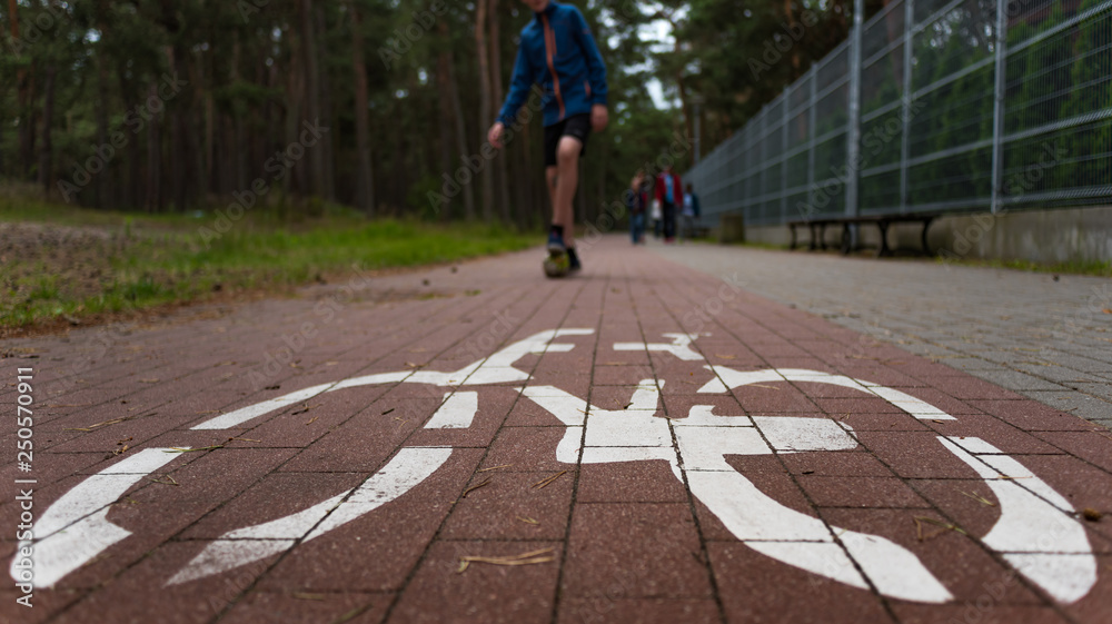 A wide angle view of a cycling sign drawn on the bike way in the park, with a caucasian teenager playing mini football, and a family walking in the backrgound.