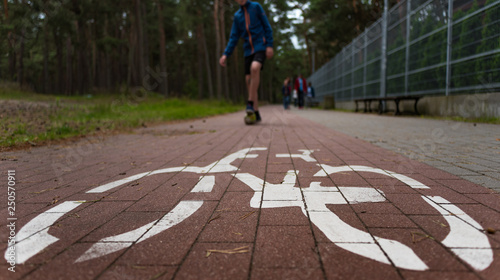 A wide angle view of a cycling sign drawn on the bike way in the park, with a caucasian teenager playing mini football, and a family walking in the backrgound.
